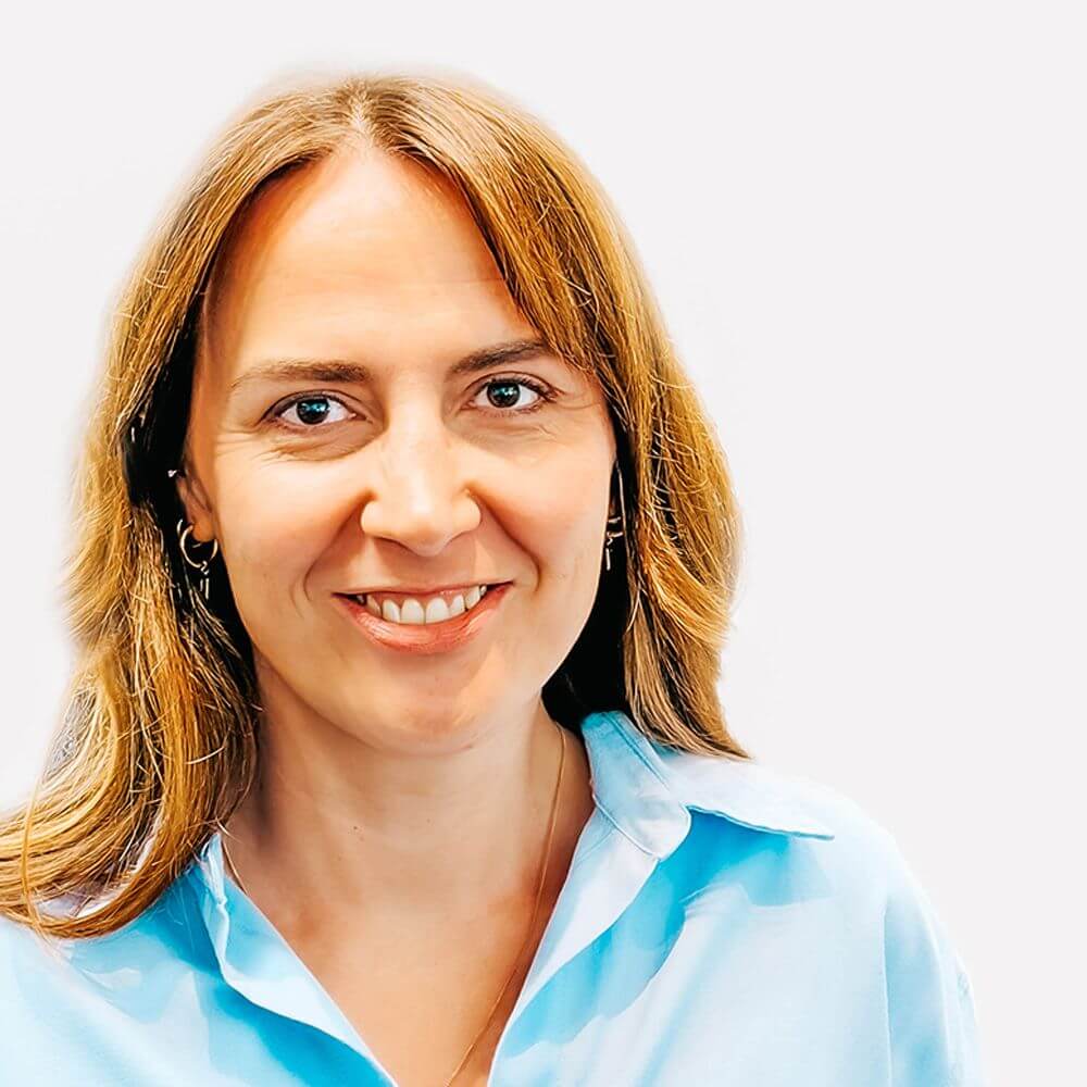 Nelli Schnee Portrait of a smiling woman with light brown hair wearing a light blue blouse, facing the camera.